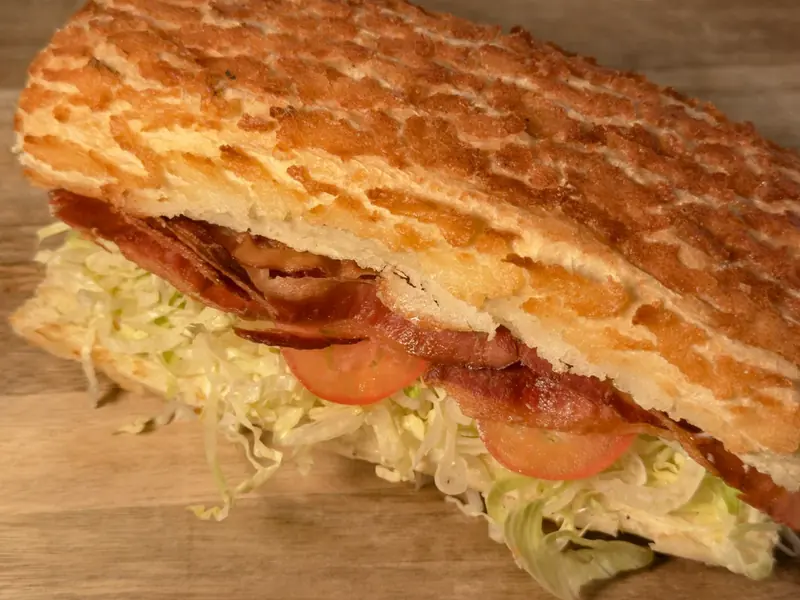 A B.L.T. cut open on a wooden board, showing crisp bacon, shredded iceberg lettuce and sliced tomato inside a griddled roll.