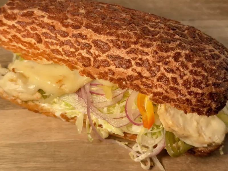 A chicken breast sandwich on a Dutch crunch roll, layered with melted cheese, shredded iceberg lettuce, red onion, sliced pepper and jalapeño.
